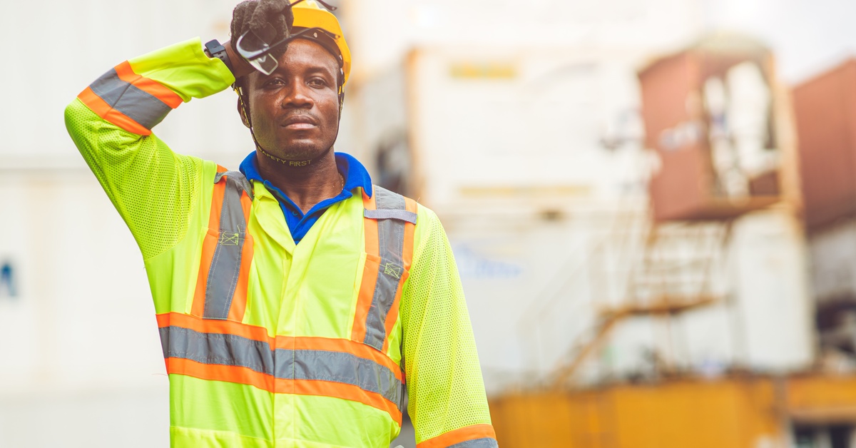 A man in a yellow reflective shirt and a yellow hard hat holds his glasses and wipes sweat from his forehead.
