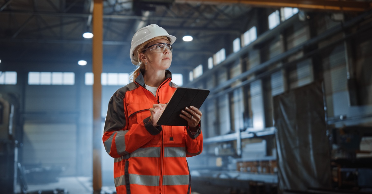 An industrial worker in an orange safety jacket and a white hard hat holds an electronic tablet and looks up.