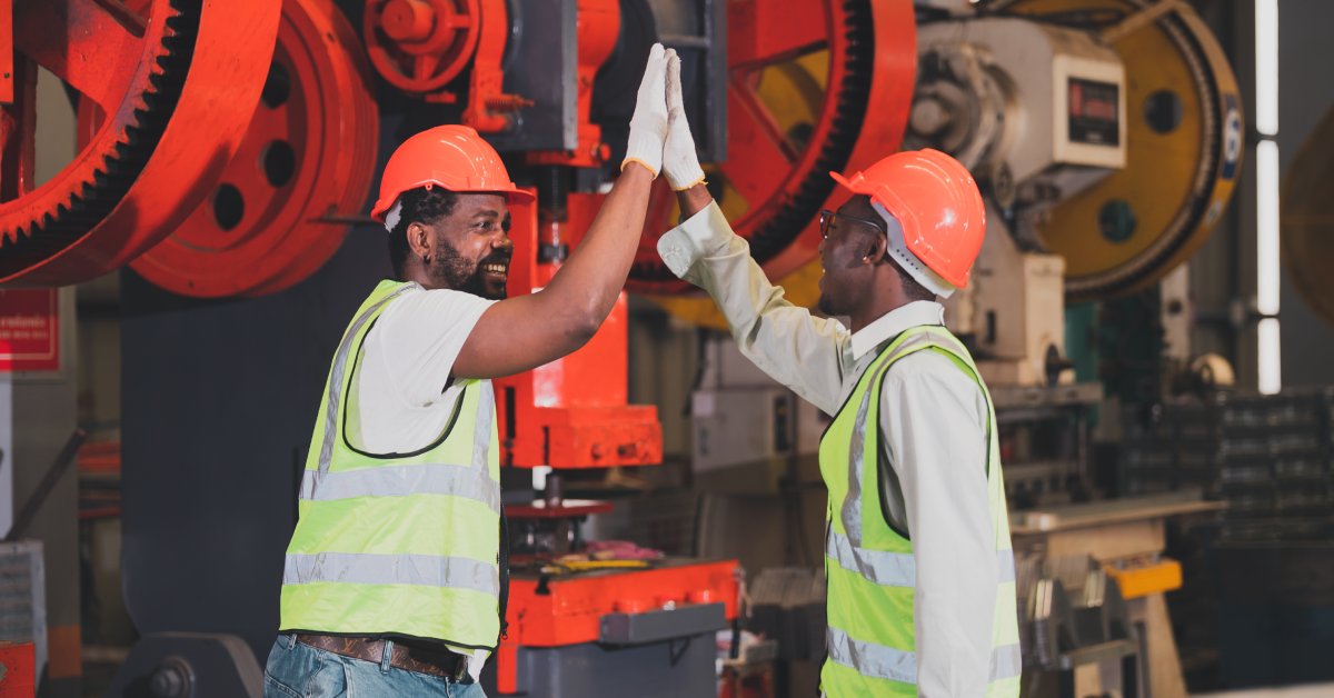 Two men working in an industrial setting and wearing yellow safety vests and orange hard hats high five each other.