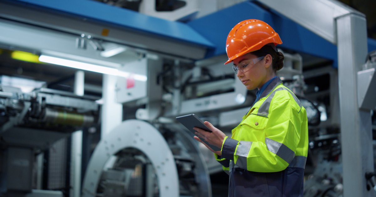A woman in a yellow work shirt, an orange hard hat, and eye protection looking down at the tablet in her hands.