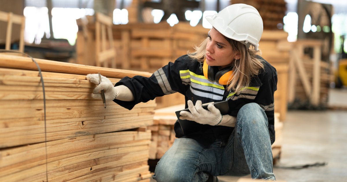 A woman in blue jeans, a dark work shirt with reflective material, and a white hard hat squatting to inspect planks of wood.