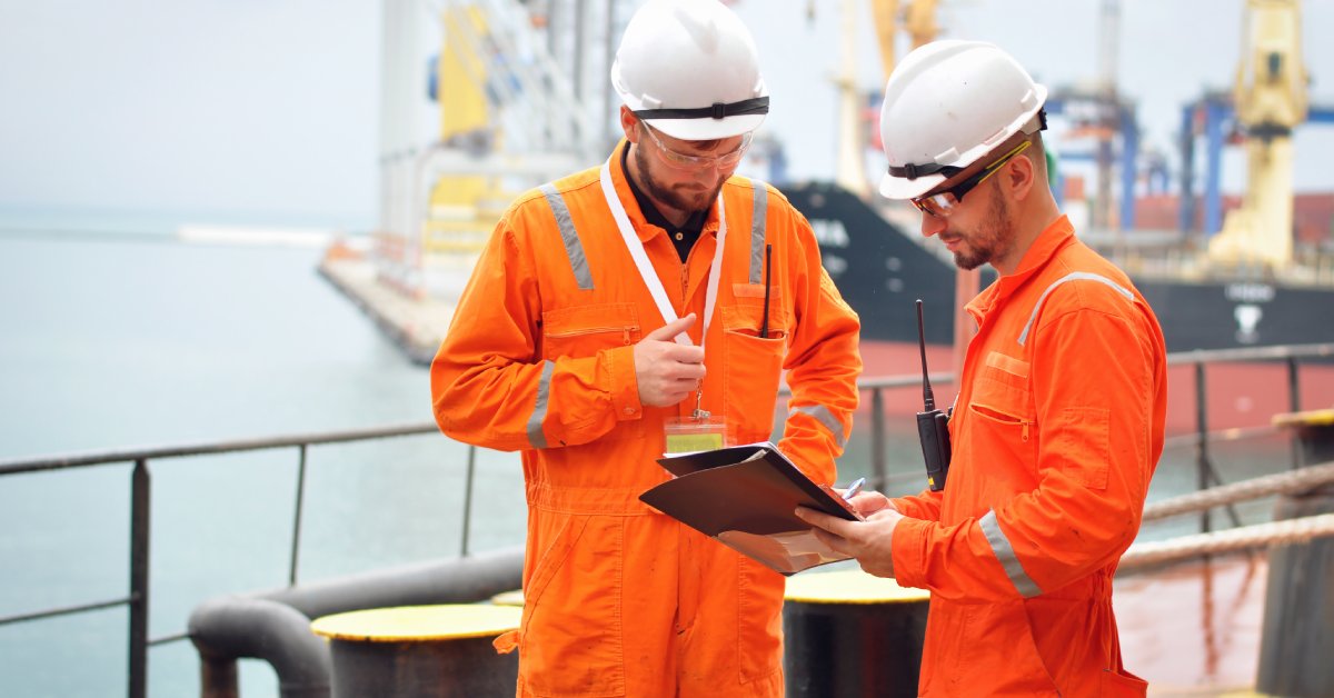 Two industrial employees in orange coveralls and white hard hats stand together and look at some papers.