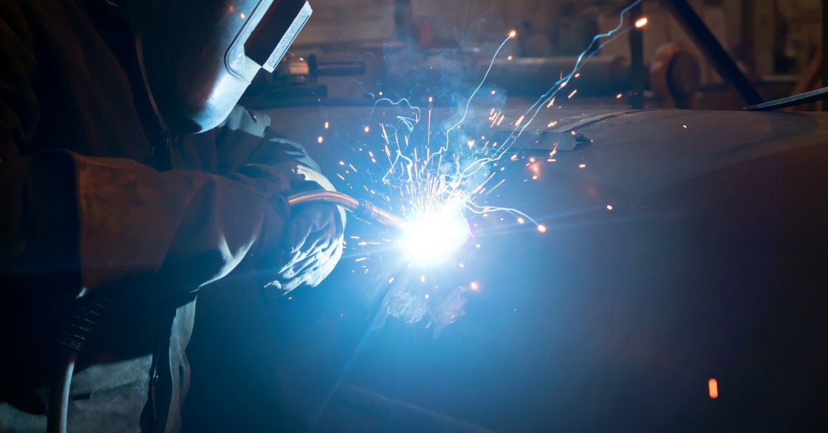 A welder in full protective clothing and a face shield uses an arc welding tool on a large structure.
