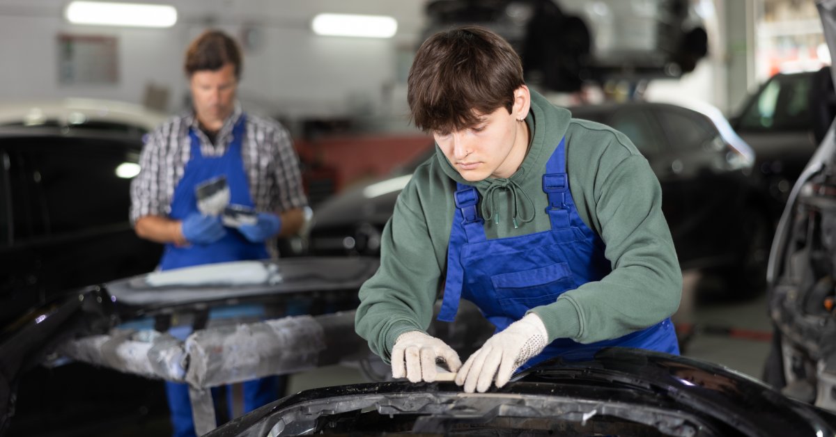 An automotive worker wearing a green hoodie under blue coveralls fixes a car in a shop with another worker behind him.