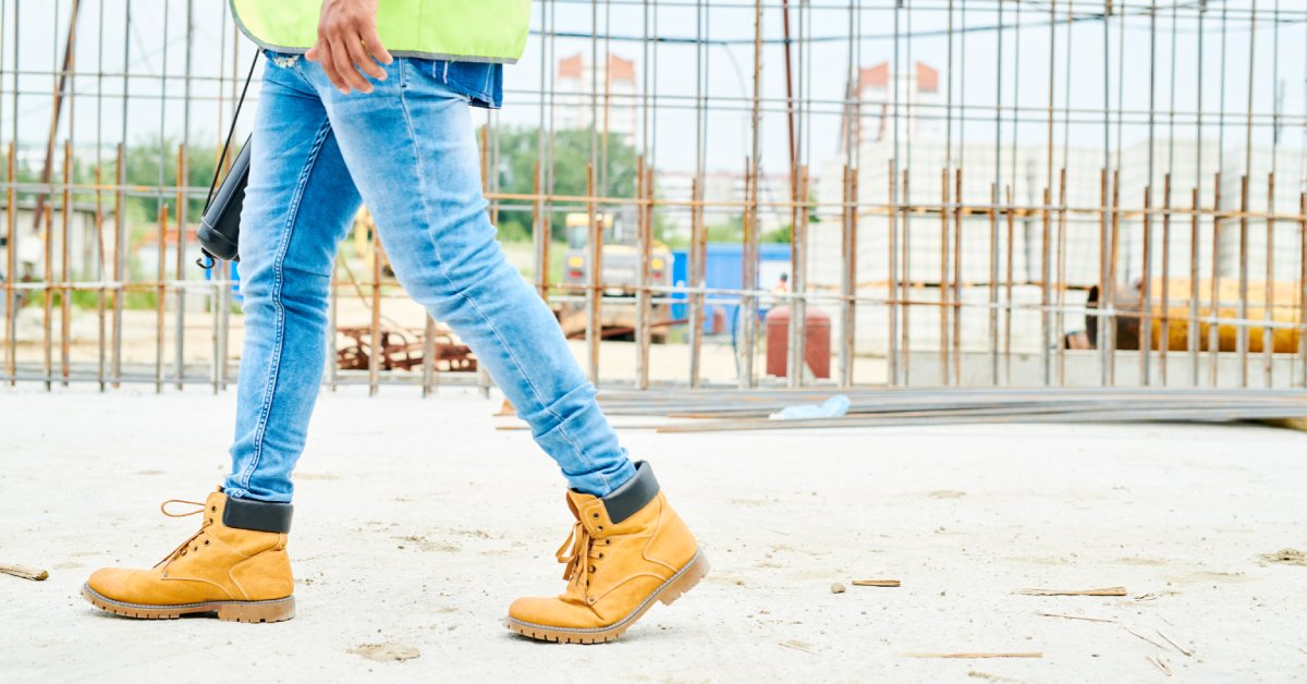 A worker in blue jeans, a yellow safety vest, and brown boots walks around at an outdoor construction site.