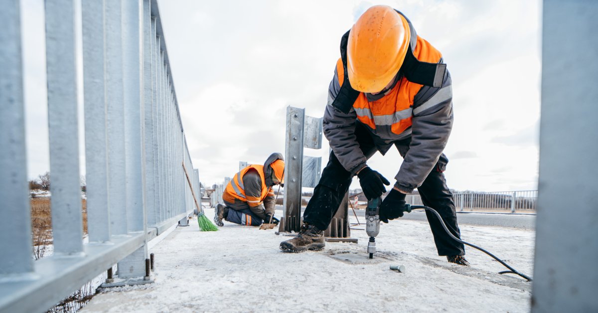 Two workers in orange high-visibility vests and hard hats do construction tasks on a road out in the snow.