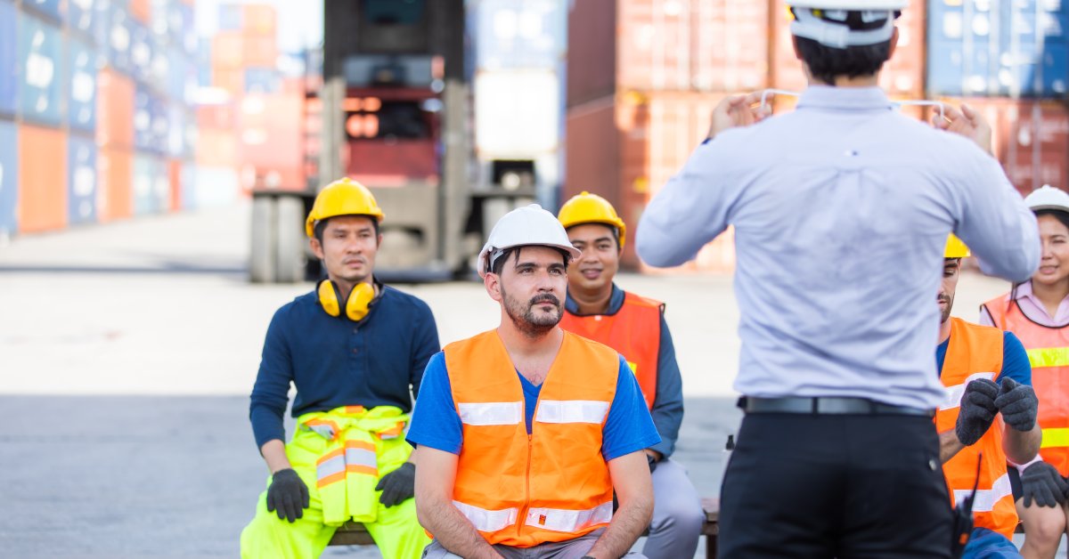 A group of workers in reflective clothing and hard hats sit and listen to a man who is standing up during a meeting outside.