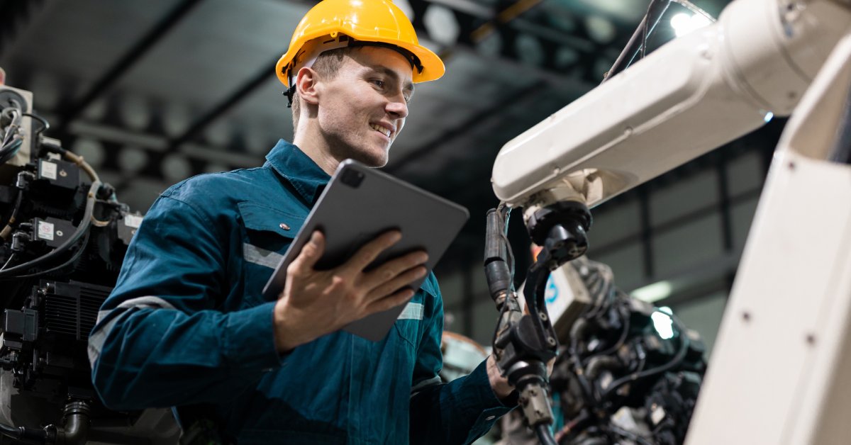 A man in blue coveralls and a yellow hard hat holds a tablet while working with a machine at an industrial job.