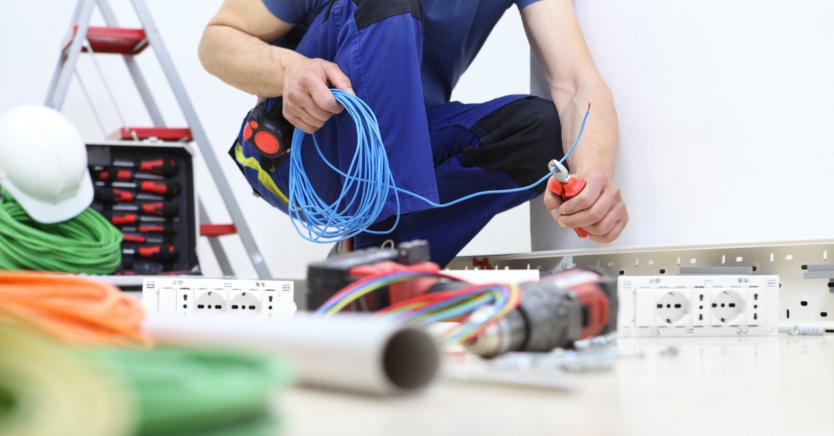 An electrical worker in a blue outfit crouches and holds a blue wire above a floor cluttered with tools.