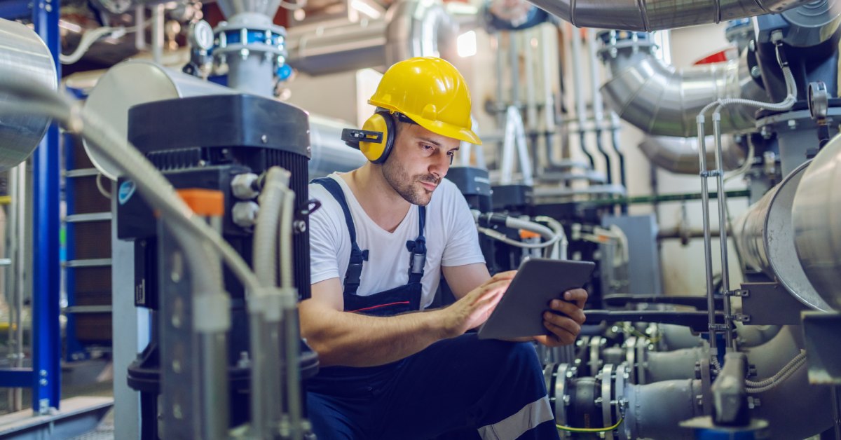 A man in a white T-shirt, blue overalls, and a yellow hard hat crouches and uses an electronic tablet in a factory.