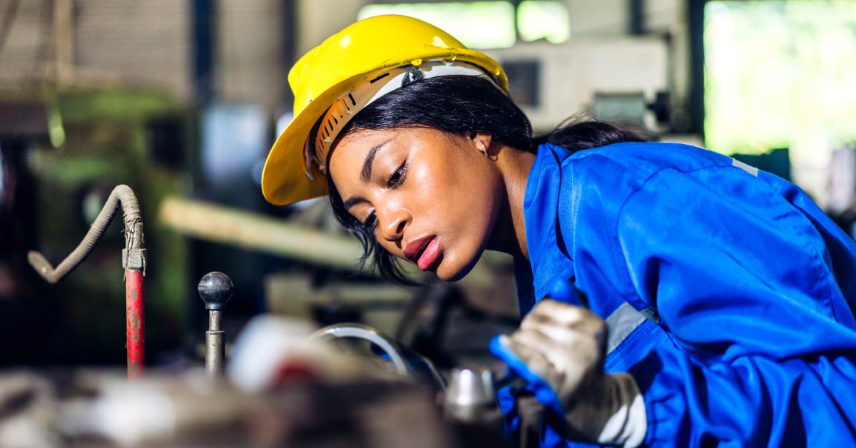 A woman with her hair in a ponytail under a yellow hard hat wears blue industrial clothing while working.