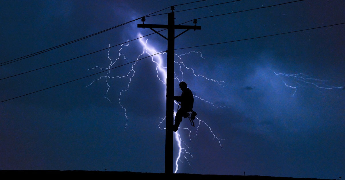The silhouette of a lineman working on the electrical pole shows up in front of a lighting bolt in a stormy sky.