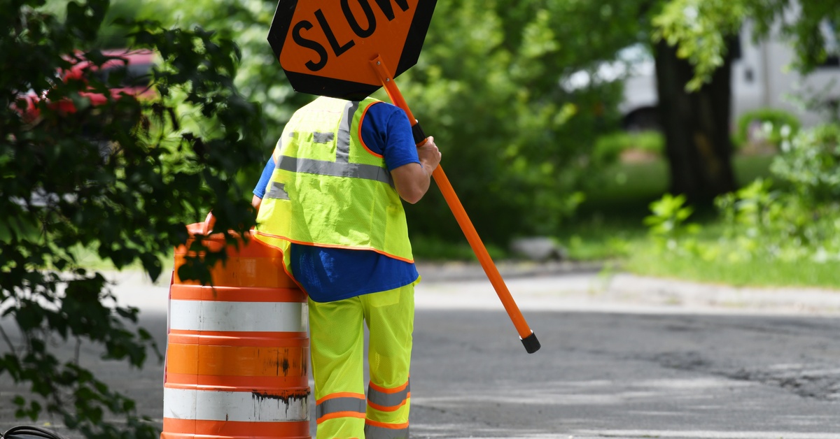 A road construction worker in yellow pants and safety vest walks forward with an orange cone and sign that says 'SLOW.'