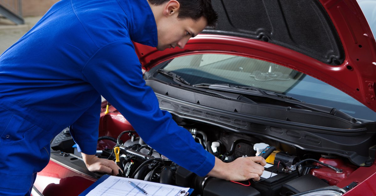 A man in blue coveralls leaning forward to look inside the hood of a red car. There is a blue clipboard and pen resting near the car's engine.