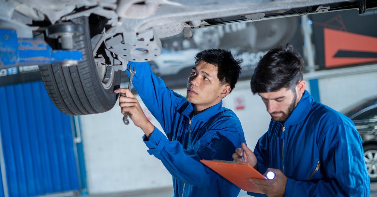 Two men in blue coveralls standing under a car. One man is holding a clipboard and the other is using a wrench on the car's tire.