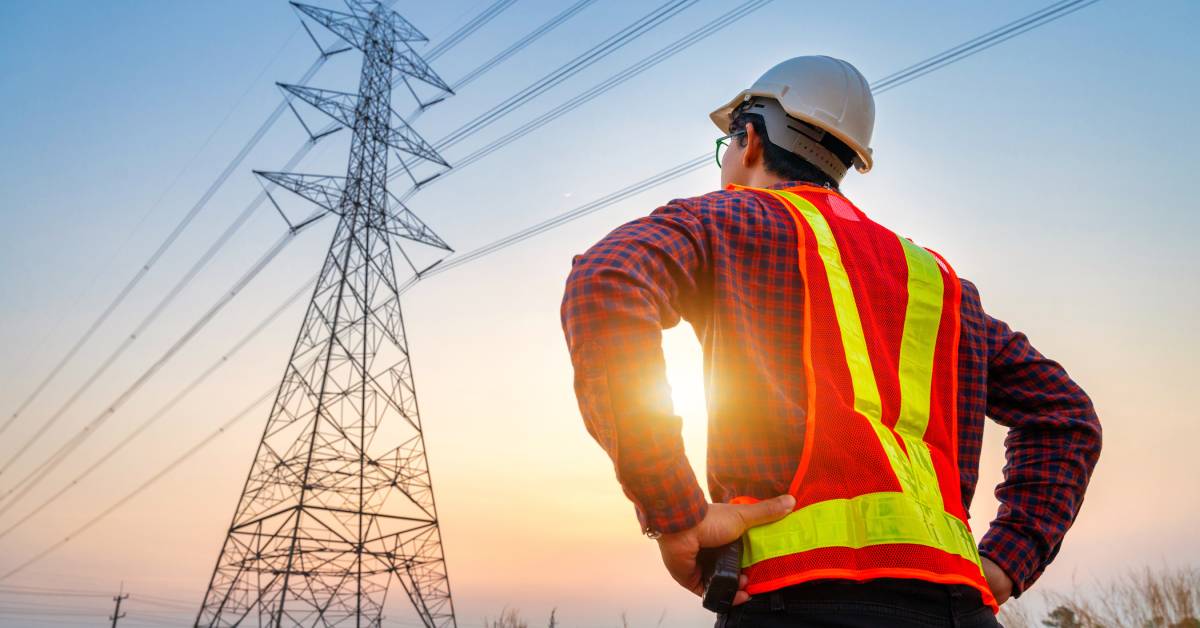 A man with an orange safety vest and a white hard hat looks up at an electrical tower during the sunset.