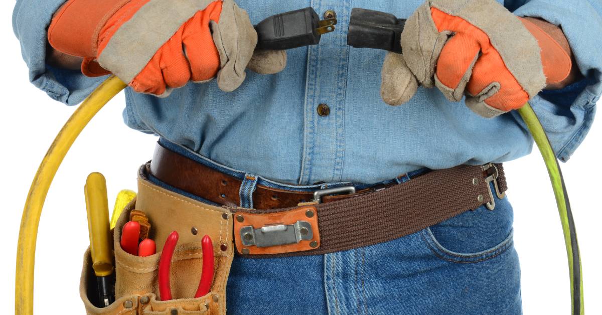 An electrician wearing a blue shirt, jeans, orange gloves, and a brown belt holding his tools plugs in a cable.