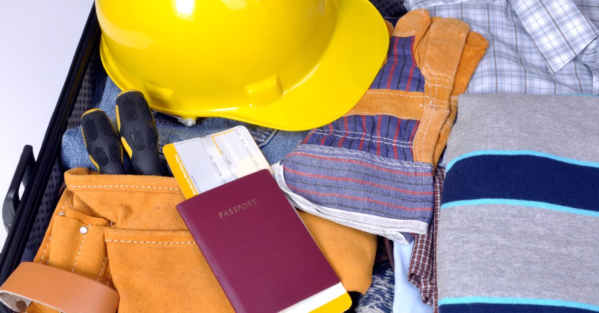 A suitcase sits open and packed with clothes, work gloves, a passport, a tool belt, and a yellow hard hat.