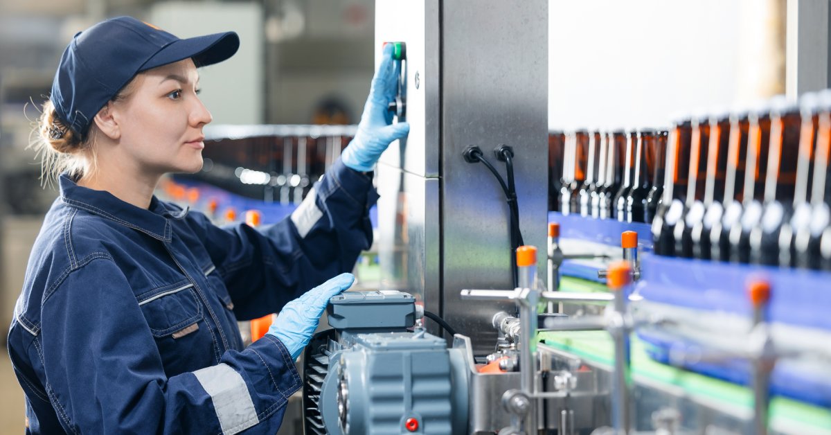 A woman working an industrial job in blue coveralls and a hat operates a piece of machinery in the factory.
