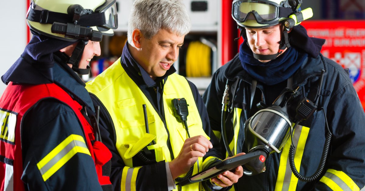 Three people in firefighter protective gear with radios on their chests stand huddled around a tablet. 