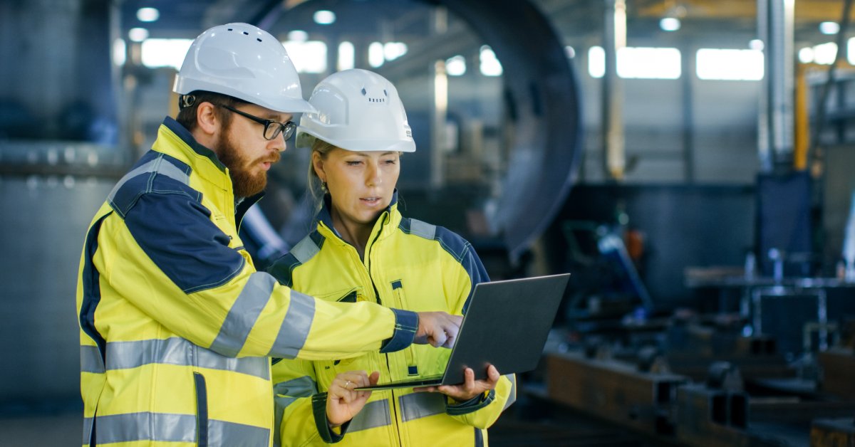 A male and a female industrial employee in yellow work shirts and white hard hats, standing together and looking at a laptop.