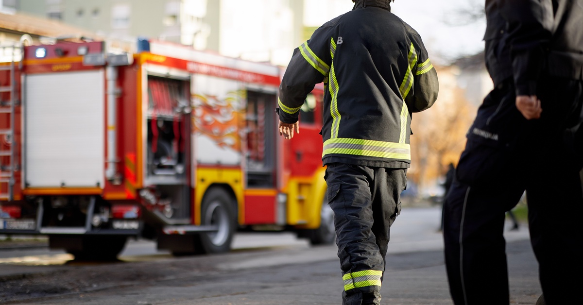 A firefighter is walking towards a firetruck. They are outside and it is daylight. The firetruck is blurry in the background.