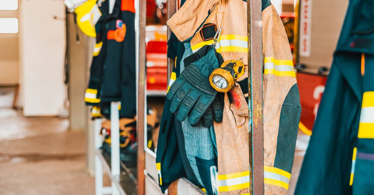 Various different types of fire resistant clothing hanging on wooden racks in a firehouse during the day. 
