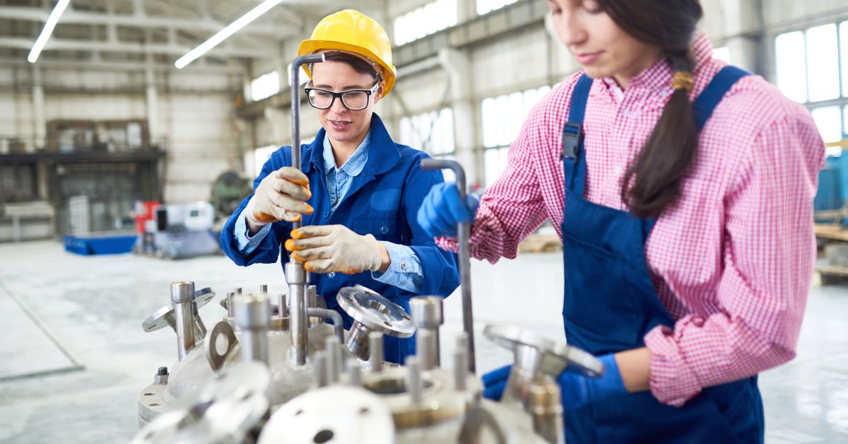 An industrial worker in blue overalls and one in blue coveralls work together on a project made of metal.