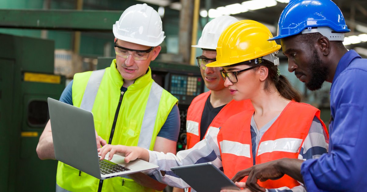 Four employees in high-visibility vests and hard hats gather together to look at a laptop and tablet.