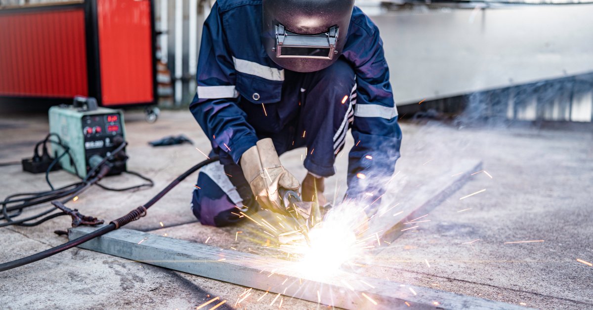 A metalworking industrial worker in blue coveralls makes sparks fly while working on a piece of metal on the ground.