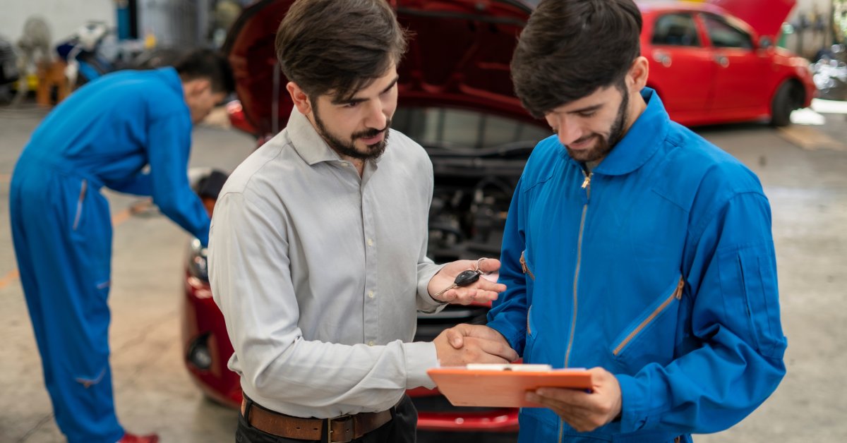  A car mechanic in blue coveralls holding a clipboard talking to a man in a button-up shirt holding a car key.