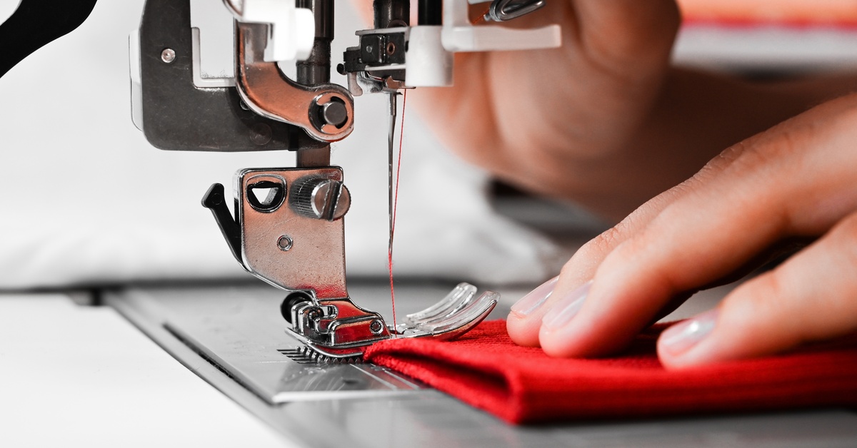 A pair of hands carefully guide the red fabric of a piece of clothing through a sewing machine using red thread.