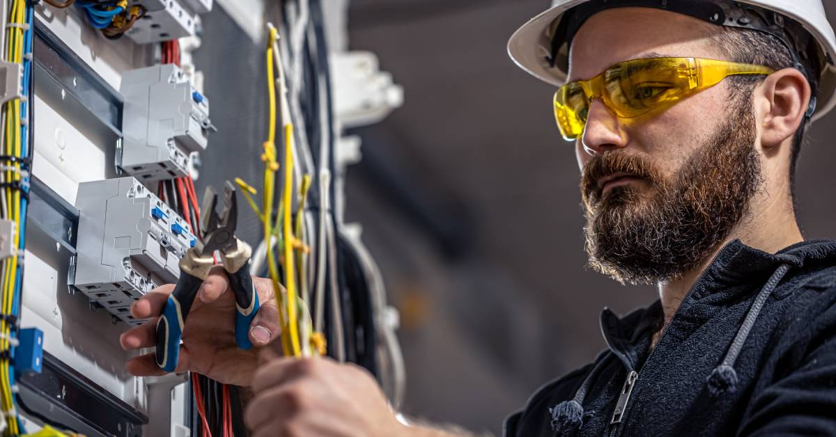 A bearded man wearing a white hard hat and yellow safety glasses uses pliers to cut wires while working.