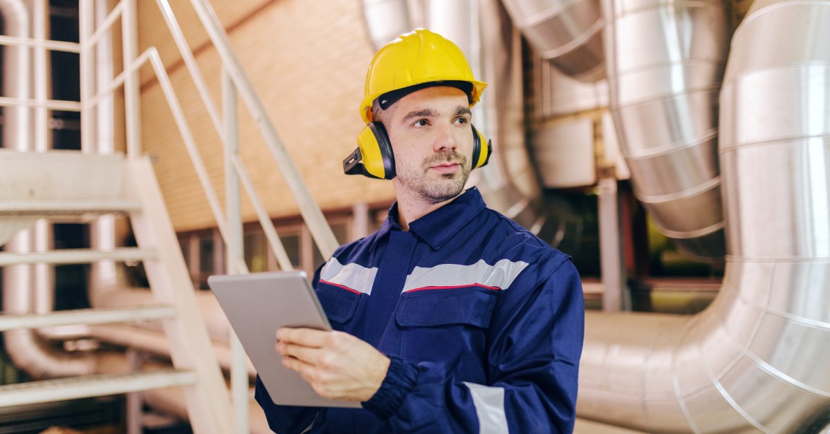 A man in an industrial workplace uniform with a hard hat and hearing protection holds a tablet and looks to the side.