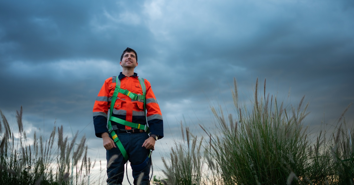 A man in industrial flame-resistant work clothes and a safety harness stands outside under gray storm clouds.