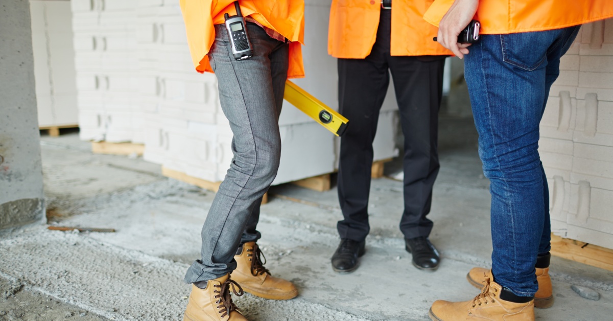 Three workers in orange vests stand together at a jobsite. Two of them are in jeans and boots, and the other has dress shoes.