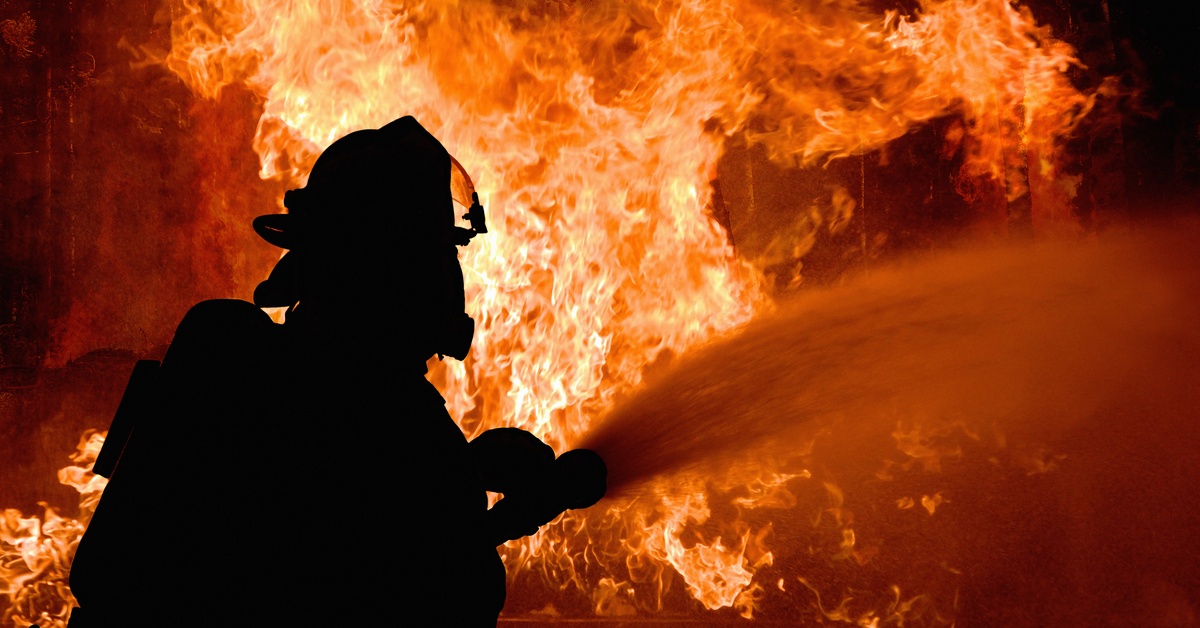 A person is holding a firehose and is using it against a flame. The person is in silhouette and has a helmet on.