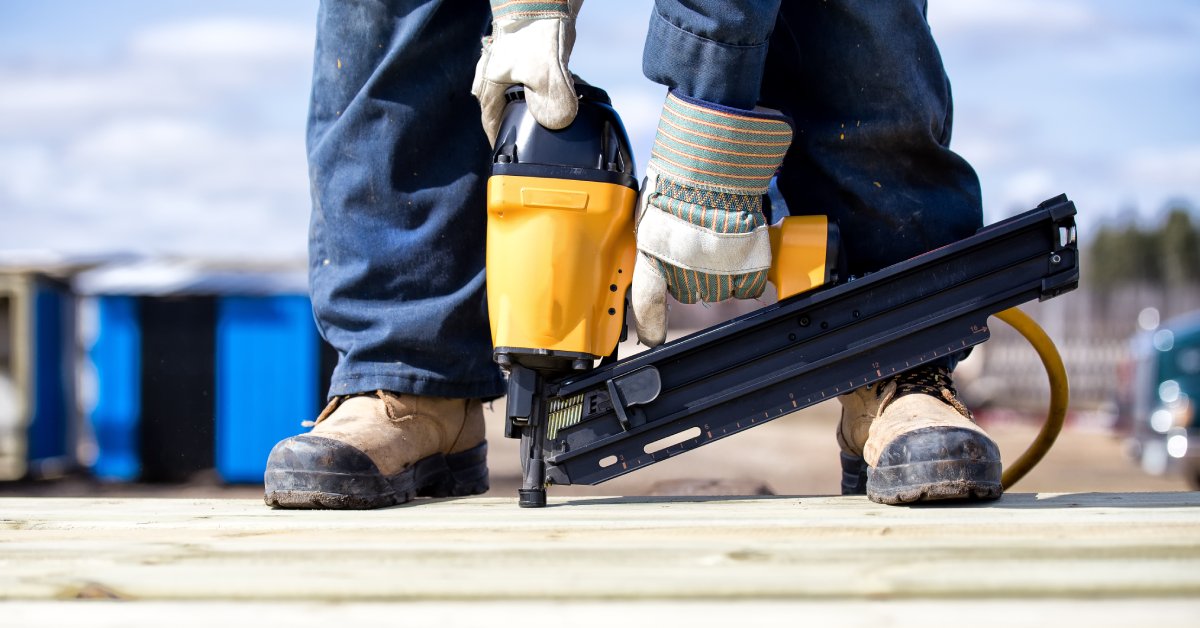 A person wearing gloves and steel toe boots. They are bending down and holding a nail gun.