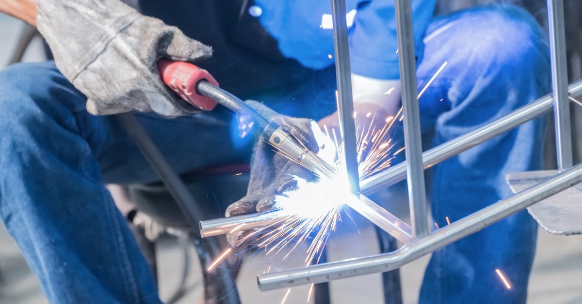 A welder in blue clothes sits on a chair with their knees spread and uses a welding tool on a piece of metal.