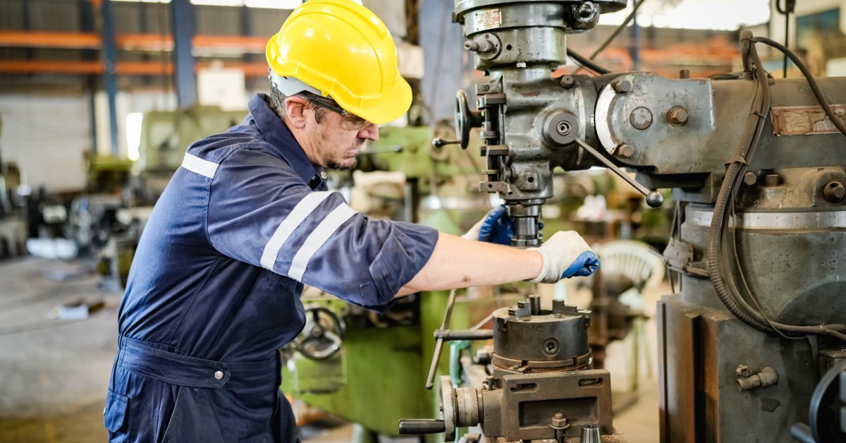 An industrial worker in blue coveralls, blue gloves, and a yellow hard hat works on a piece of machinery.