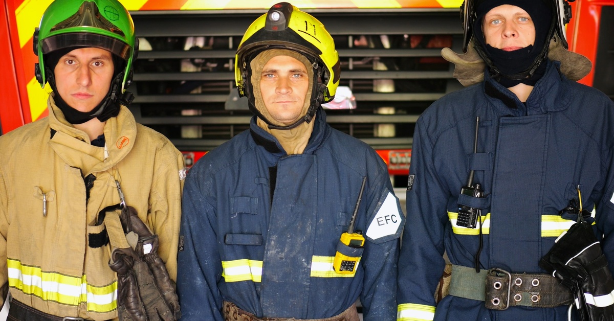 Three firefighters dressed in their flame resistant uniform with helmets and fabric head accessories stand together.