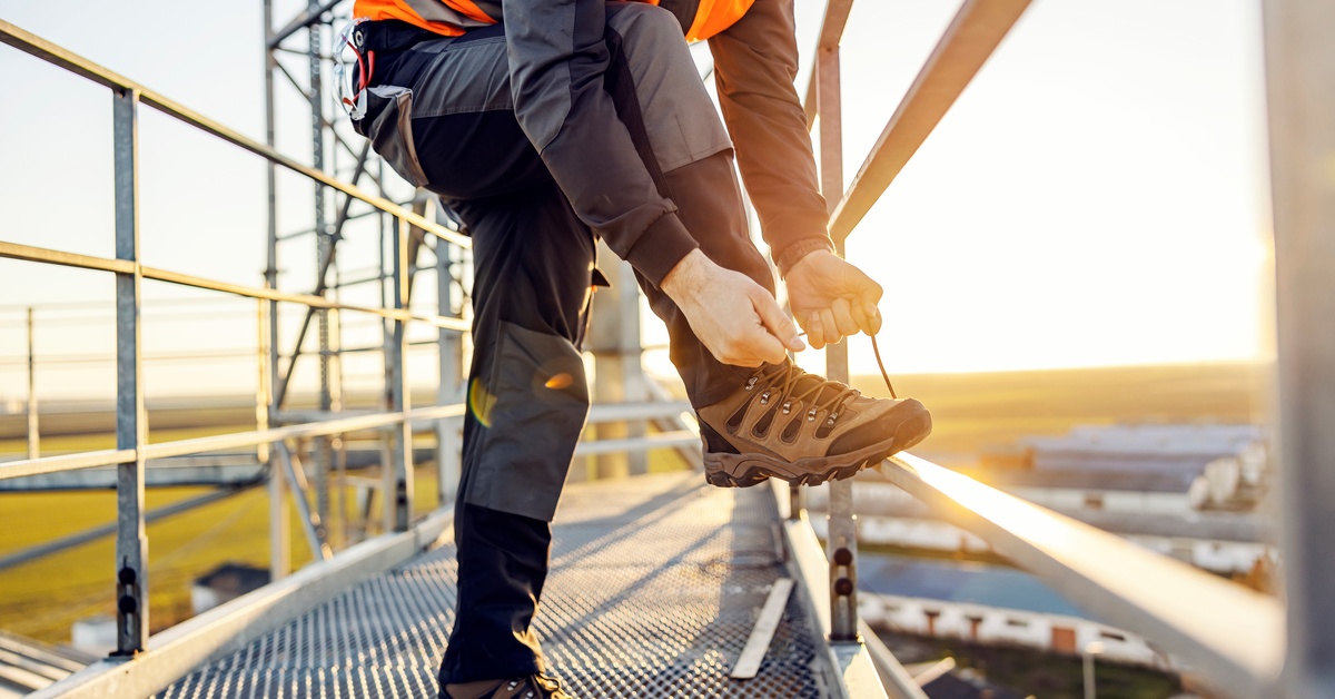 An industrial worker in an orange vest holds up his foot to tie his brown work shoe while on a scaffold.