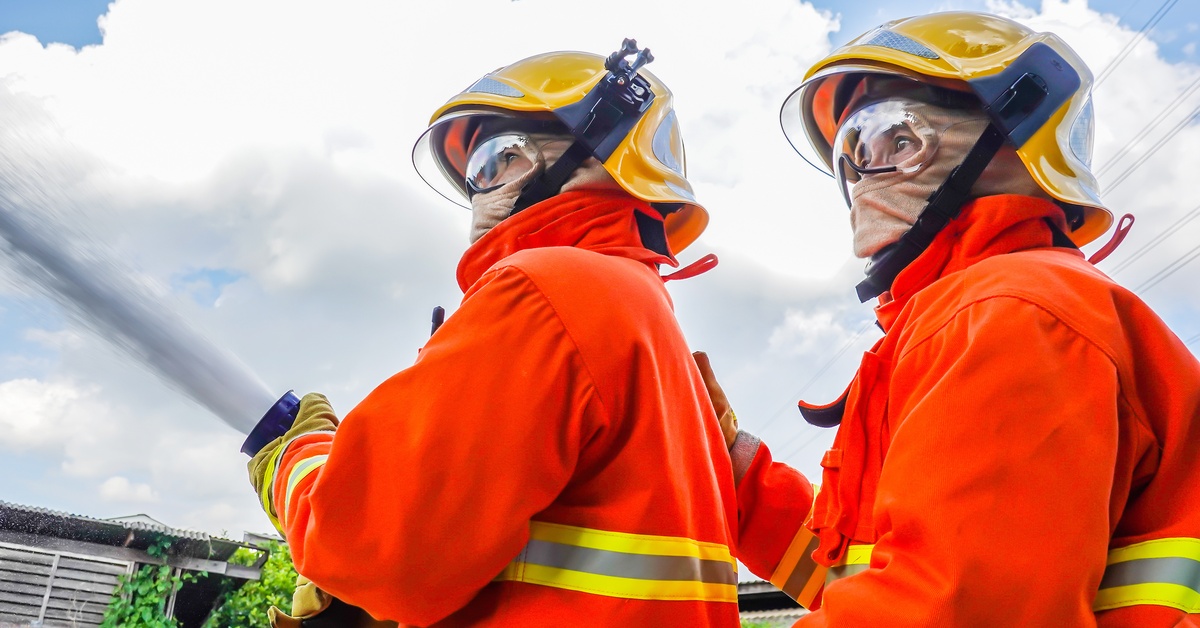 Two firefighters in visible orange work clothes and helmets stand outside with a large pressurized water hose.