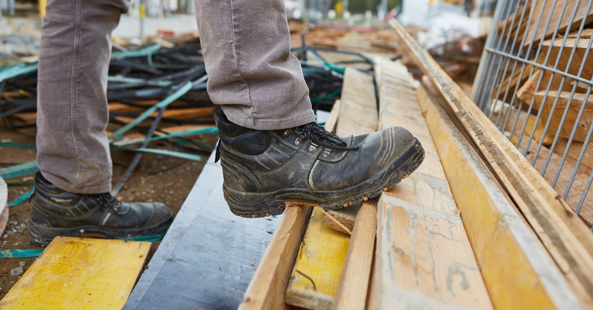 A worker wearing black steel-toe boots presses the sole of one into a pile of wood on the floor of a construction site.