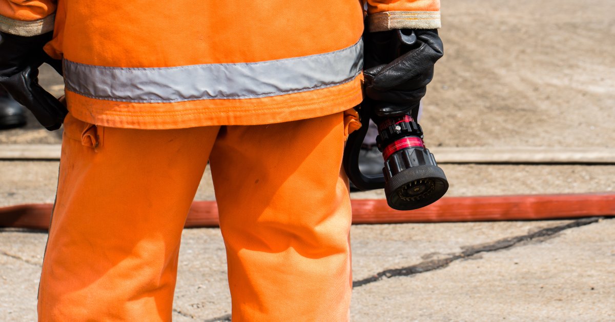 A fireman in an orange flame-resistant protective outfit and black gloves holds a water hose by the nozzle.