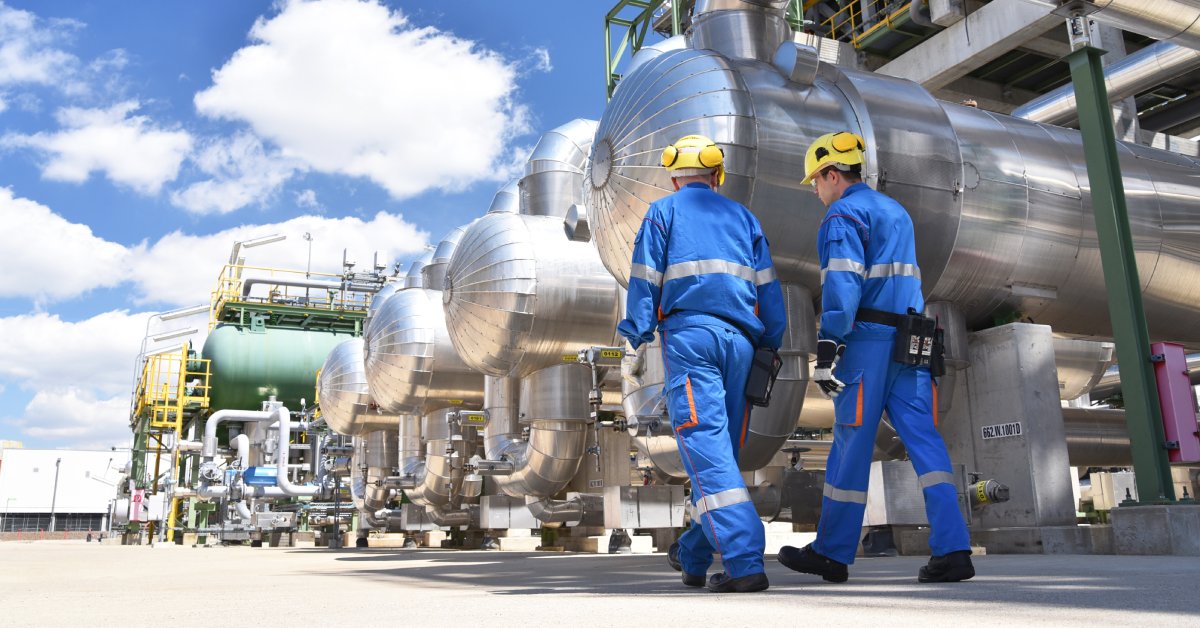 Two industrial workers in full blue protective clothing and hard hats walking outside next to metal containers.