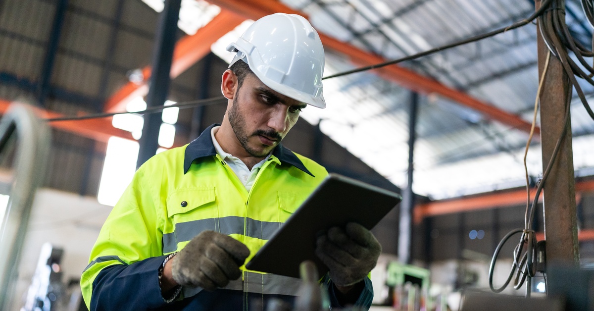 An industrial worker in flame-resistant clothing and a hard hat holds and uses an electronic tablet.