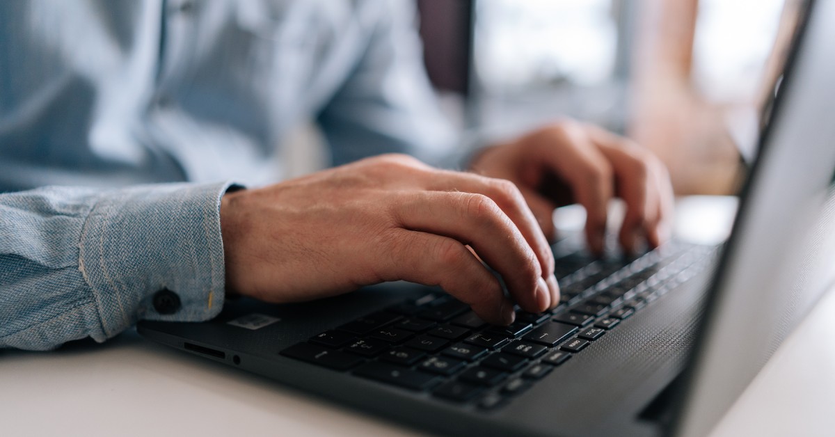 A person wearing a long-sleeve shirt types on the keyboard of a black laptop sitting on a white table.