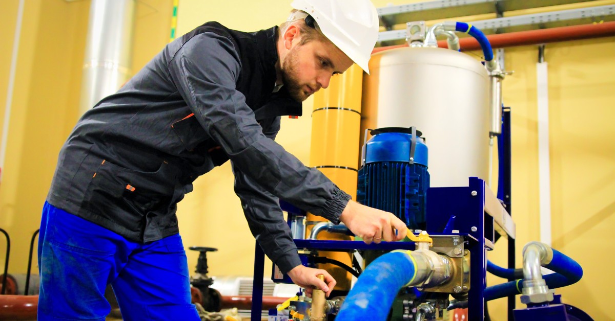 A man leans forward to work on a hose system while wearing blue pants, a gray jacket, and a white hard hat.