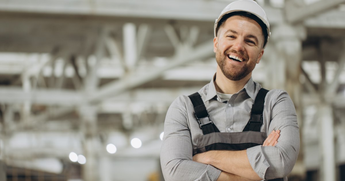 A man wearing overalls and a white hard hat stands with his arms crossed and laughs with a wide smile.